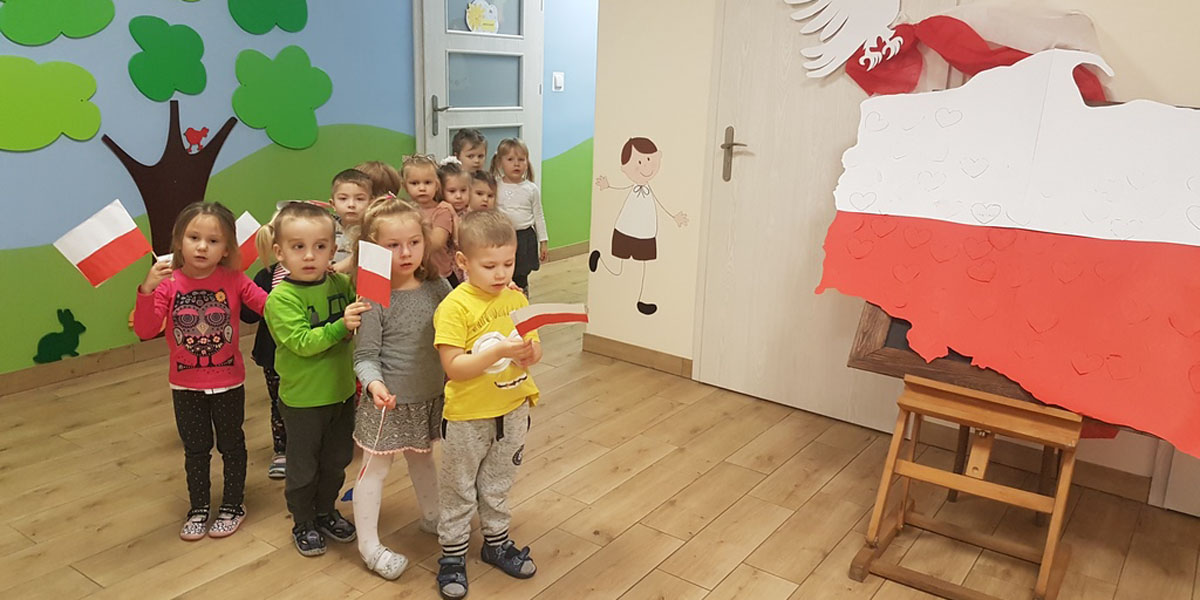 Dzieci z grupy Liski wychodzą z sali na korytarz z małymi flagami w ręce