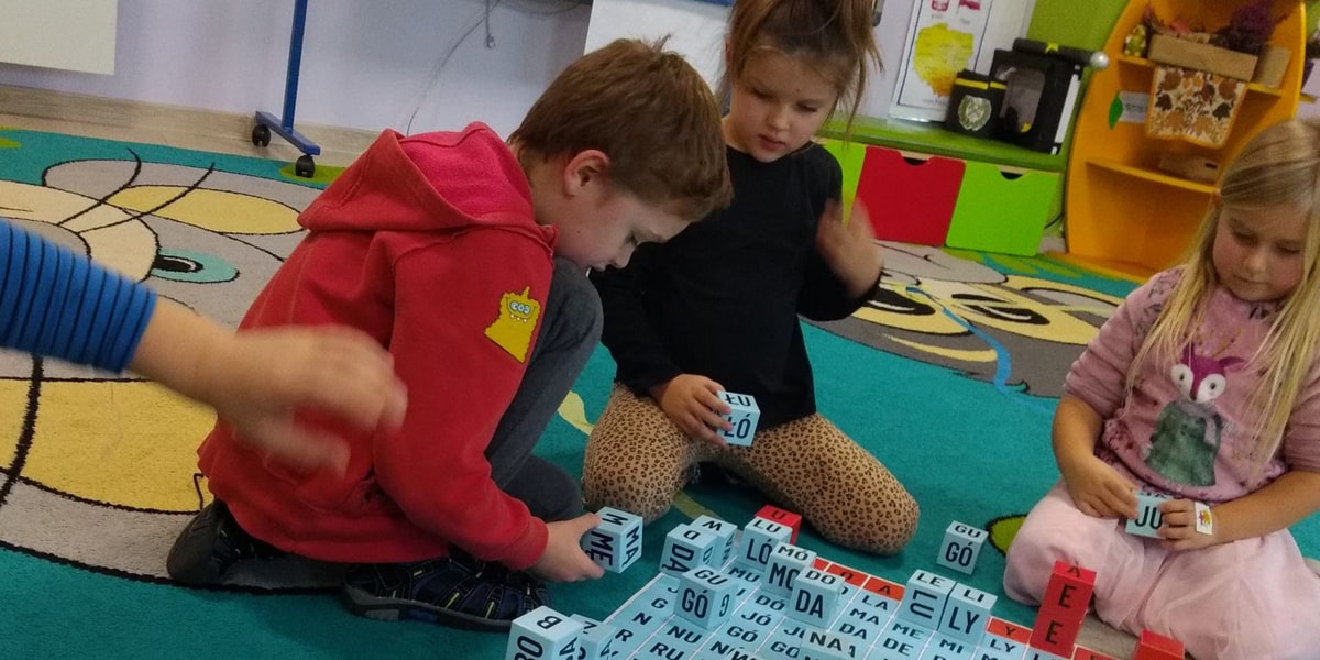 Dzieci siedząc na dywanie układają klocki z sylabami na macie do kodowania