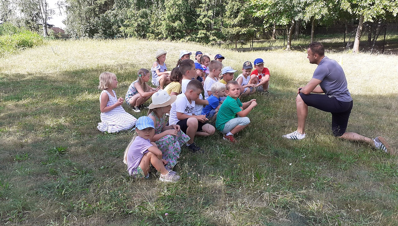 dzieci siedzą na trawie i słuchają wyjaśnień trenera jak grać w piłkę.