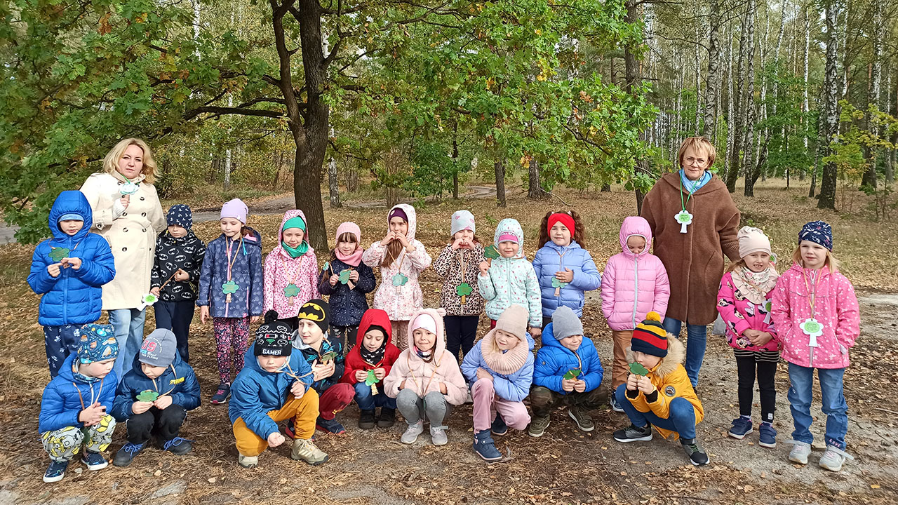 Cała grupa dzieci wraz z paniami stoi w lesie pod dębem, z zawieszonymi na szyi emblematami drzew.